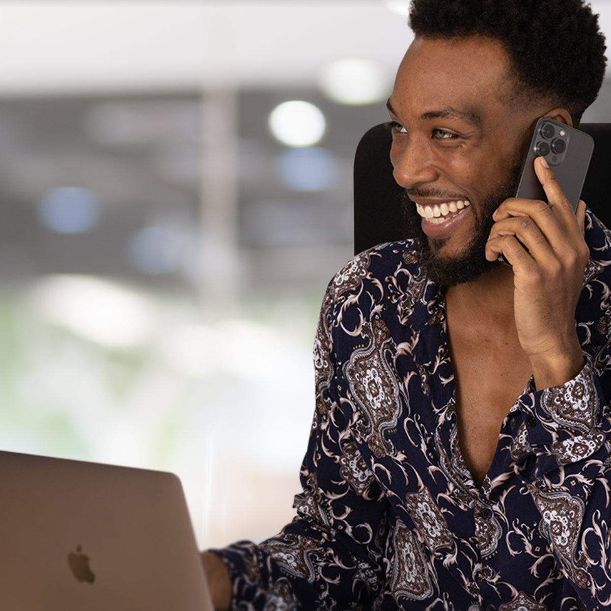 Smiling man at his desk on the phone wearing a shirt with a couple buttons undone and an invisible undershirt underneath