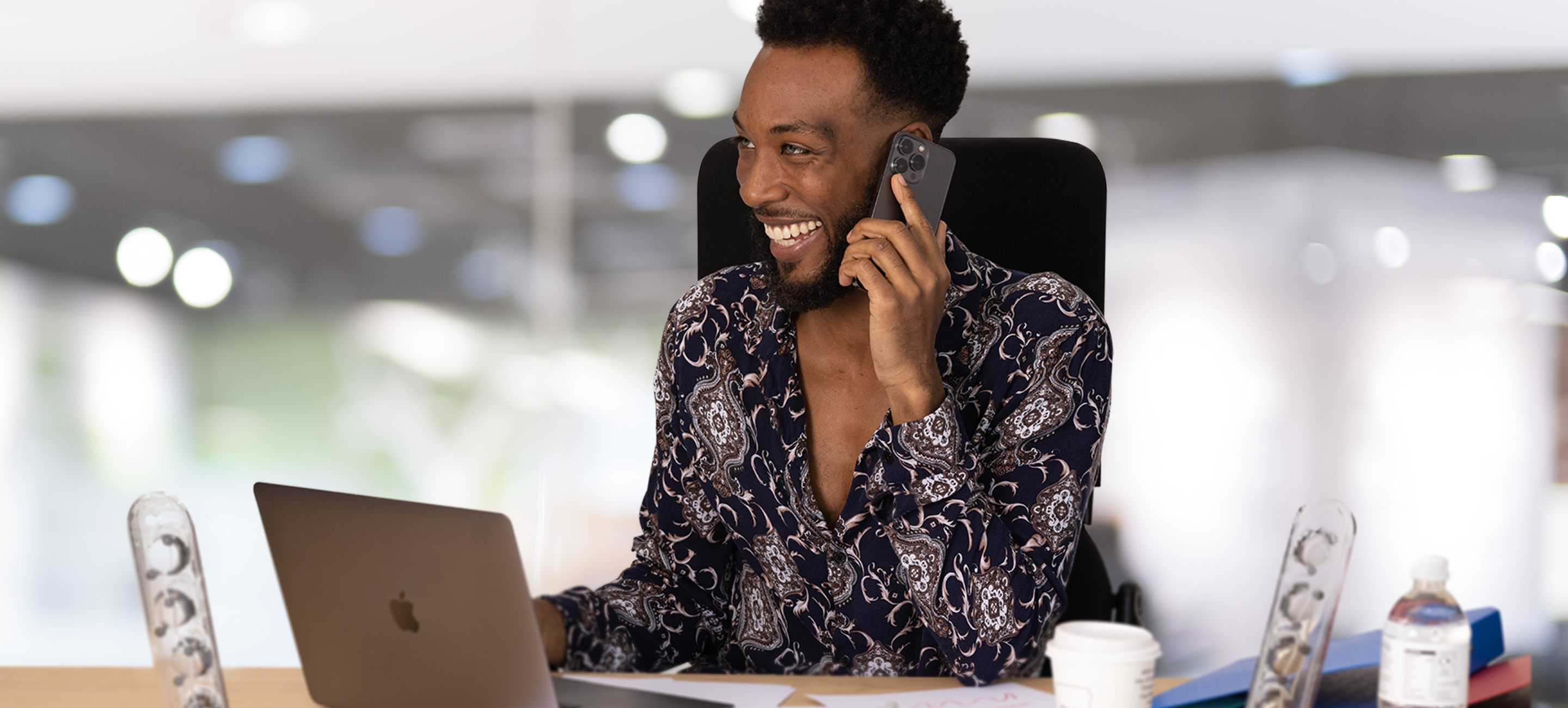 Smiling man at his desk on the phone wearing a shirt with a couple buttons undone and an invisible undershirt underneath